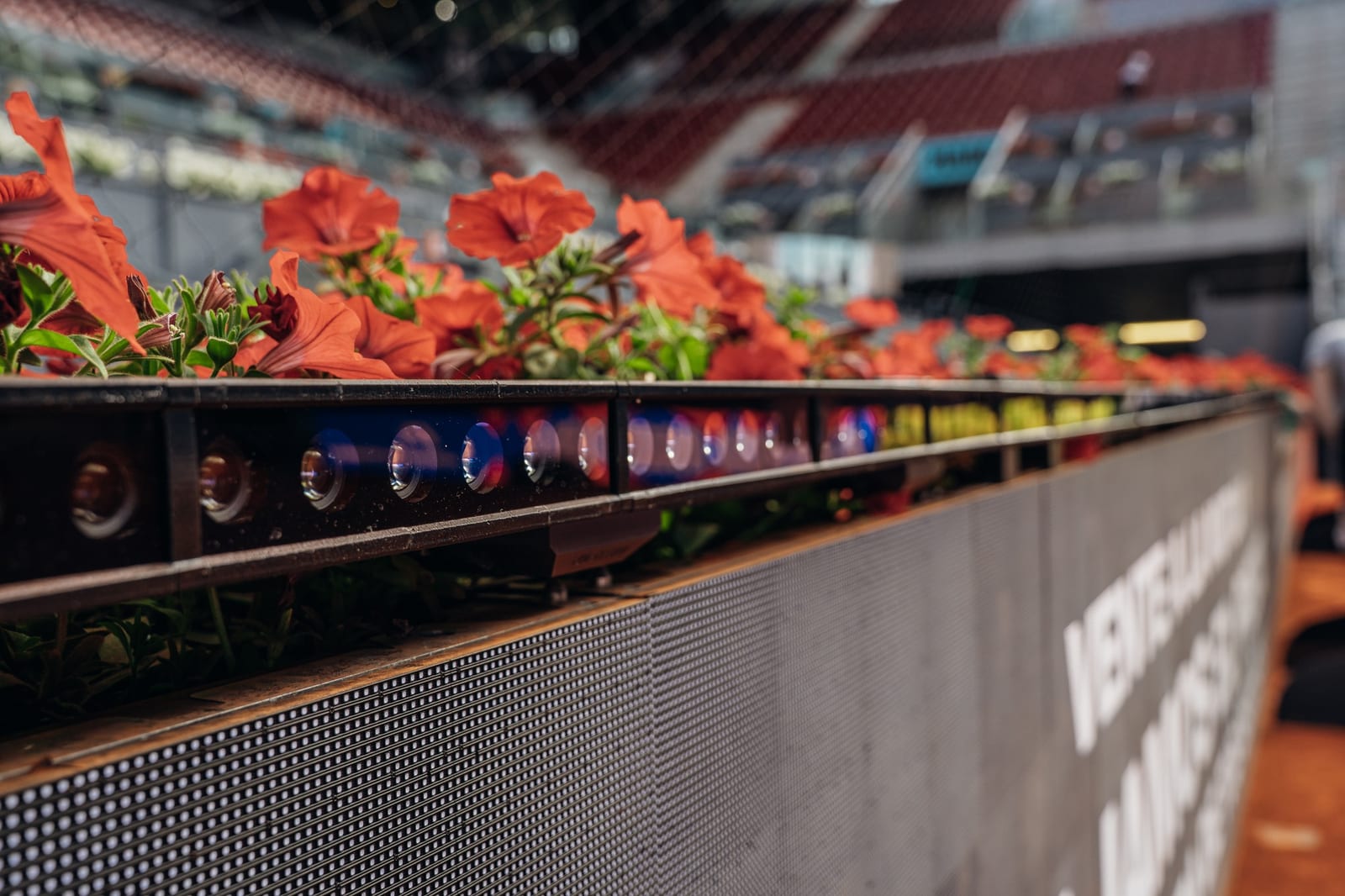 Close-up of a row of small camera lenses in a black bar set among red flowers at the edge of a tennis arena
